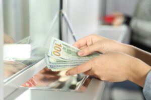 Hands holding several one hundred dollar bills at a bank counter with a glass partition.