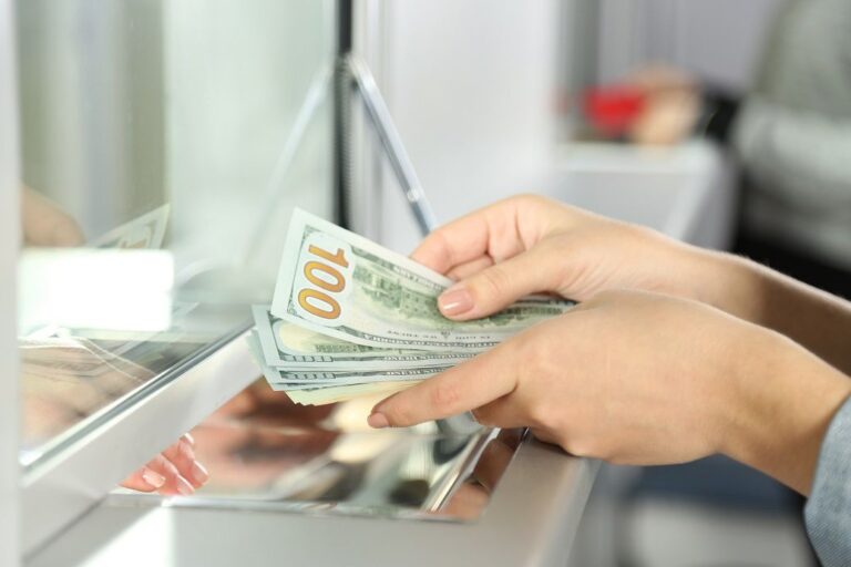 Hands holding several one hundred dollar bills at a bank counter with a glass partition.