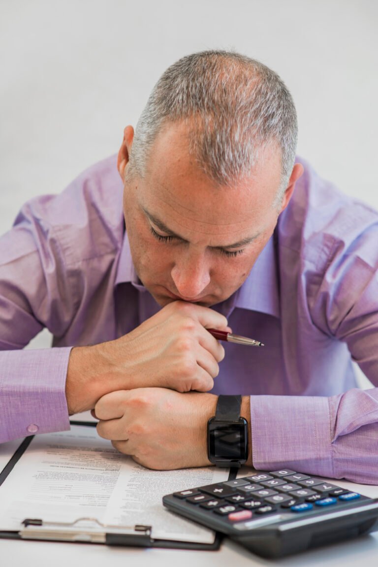 A man in a purple shirt looking down with a pen in hand, seated at a desk with documents and a calculator.